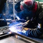 A welder works on metal fabrication indoors, creating bright sparks and smoke.